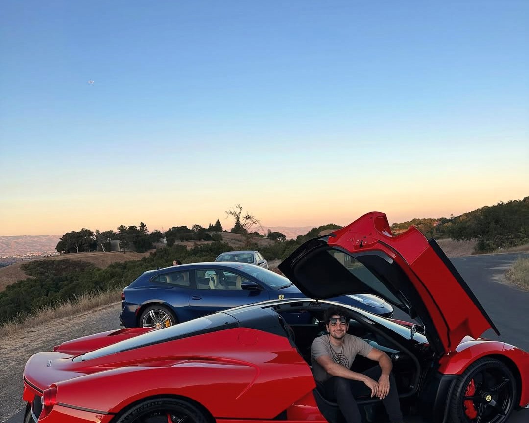 Julian seated in the open door of a red Ferrari, hills behind him