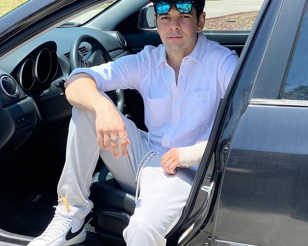 Julian in a white shirt, seated in the open door of a black car